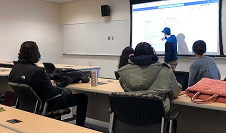 Student wearing a blue hat standing in front of class teaching fellow students sitting in class