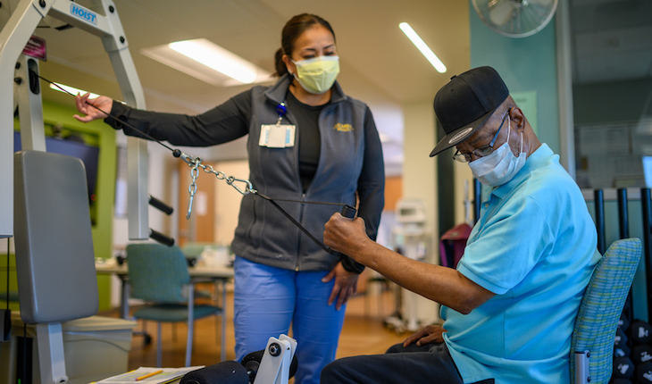 Un hombre usando una mascarilla usa un aparato de ejercicios con el brazo izquierdo mientras una asistente de salud se prepara para ayudarle.