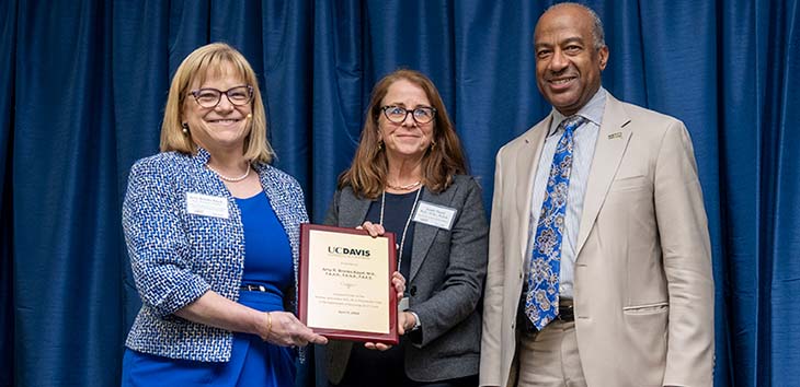 Two women and a man in business clothing are standing together smiling. The two women are holding a plaque that they point toward the camera. 