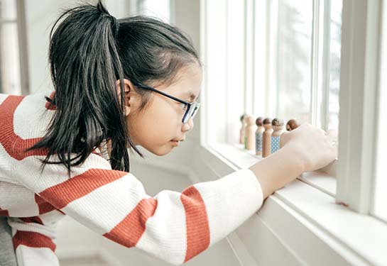 a young girl lines up wooden toys along a windowsill