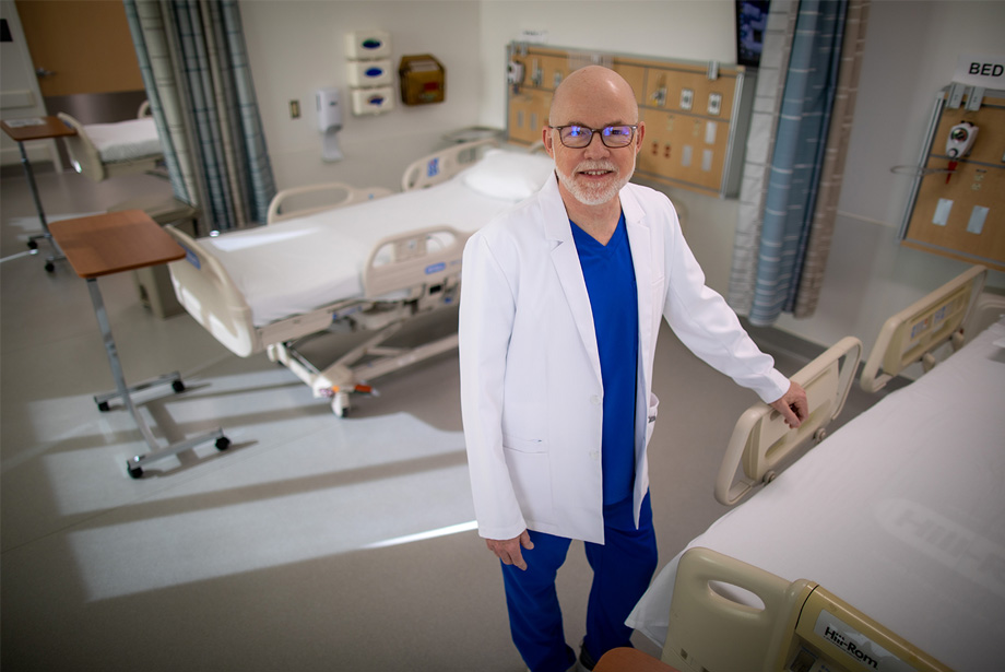 Bill Randall in scrubs and white coat stands in hospital ward