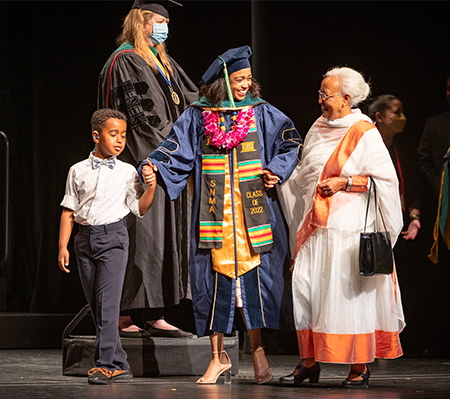 A female student in blue graduation gown holds the hand of a child and an older woman as they walk across the stage at Mondavi Center