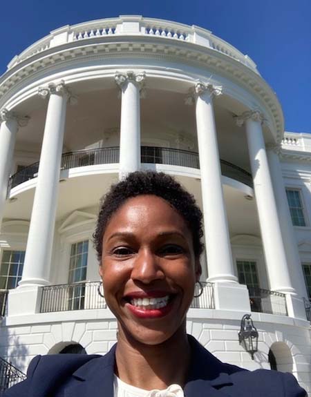 A smiling Black woman in a blue suit take a selfie with the White House portico in the background.