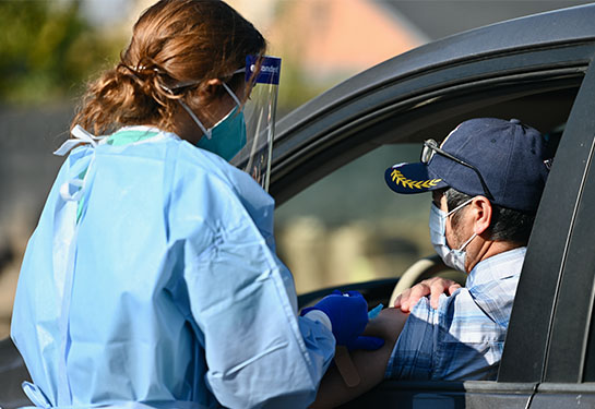A student with HLUB Clinic in a blue protective gown and face shield provides a flu shot to a man behind the wheel of his car 