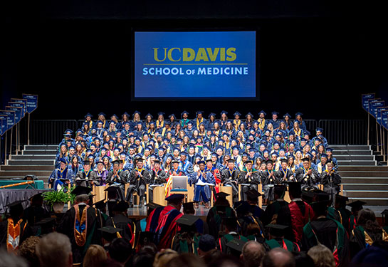 About 100 UC Davis medical students in blue caps and gowns sit on stage at the Center during their 2018 commencement
