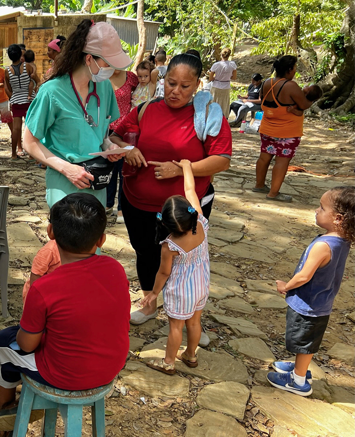 Medical student Erika Monasch in green scrubs and stethoscope around her neck speaks with a woman in an outdoor clinic in Honduras