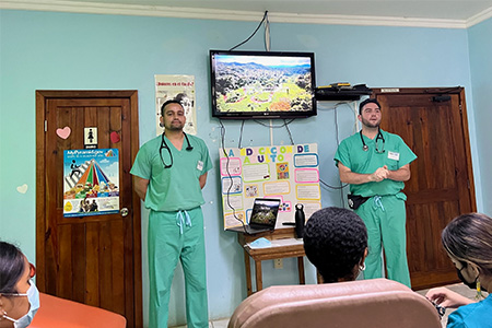 Medical students Om Patel and Brock Burnett, wearing scrubs stand before a group of adults to explain the dangers of parasites