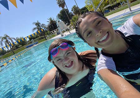 Two young women pose for a selfie in a swimming pool. One wears a swim cap and both are smiling. 