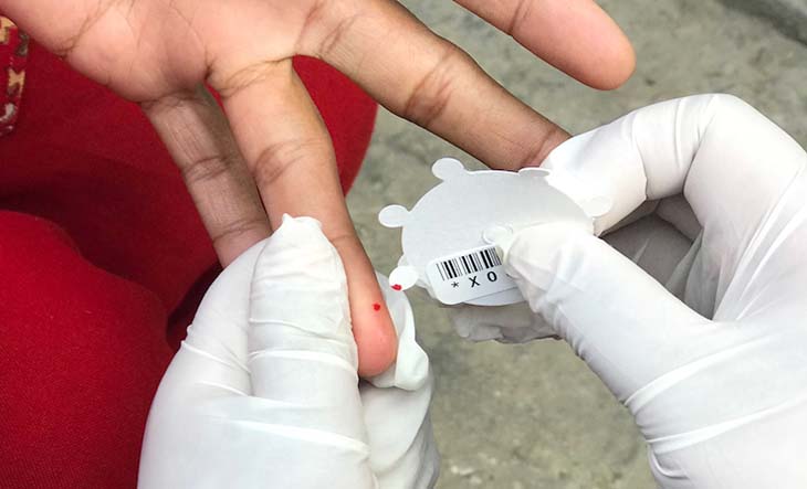 A pair of gloved hands is seen collecting a small amount of blood from a finger that has been pricked. 