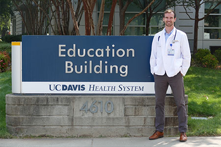 A young man in a white lab coat stands next to the sign that reads: “Education Building, UC Davis Health System”