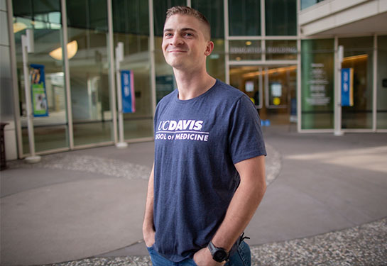  A student wearing a blue T-shirt with the words “UC Davis School of Medicine” stands in front of the school’s Education Building