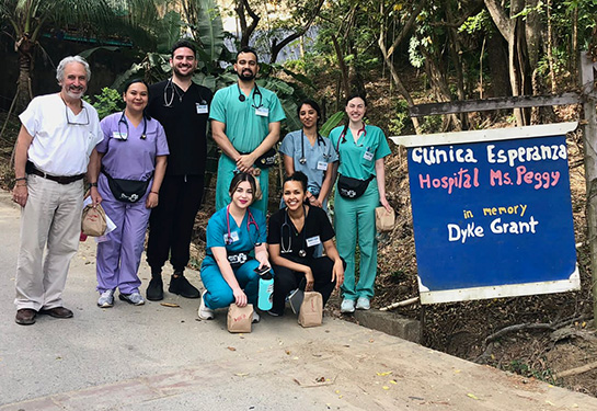 Seven UC Davis medical students wearing scrubs stand with Professor Michael Wilkes next to “Clinica Esperanza” sign in Honduras