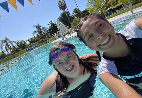 Two young women pose for a selfie in a swimming pool. One wears a swim cap and both are smiling. 