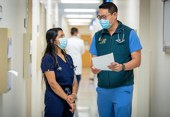 Dr. Kirti Malhotra and Dr. Joseph Kim, both in blue scrubs, stand next to each other talking in clinic hallway