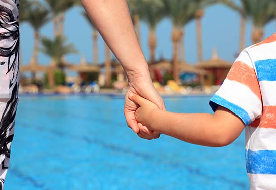 A parent holds the hand of a child standing beside a large swimming pool