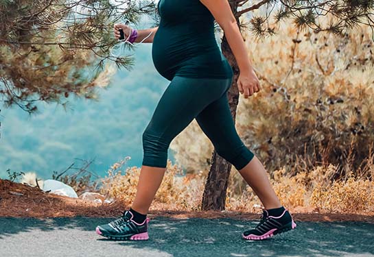 A pregnant woman walks outdoors, with pine trees in the background.