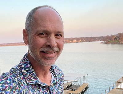 A middle-aged man in a colorful shirt takes a selfie with a lake in the background.