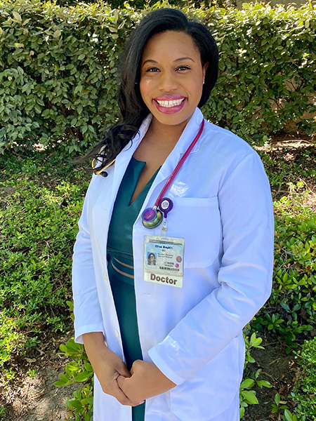 Elise Boykin-Harris wearing a white lab coat and stethoscope around her shoulders, with green shrubs in the background