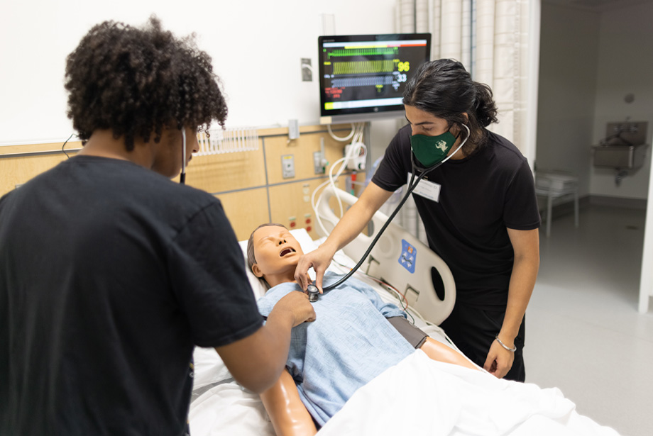 Two students stand over simulation manikin with stethoscopes