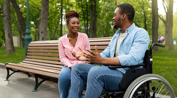 a young man in a wheelchair sits next to his girlfriend, who’s sitting on a park bench. 