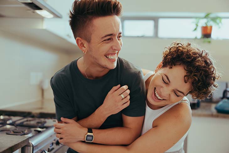 A young LGBTQIA+ couple laugh together in the kitchen