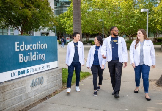 Four students wearing white lab coats walking by blue sign that says Education Building