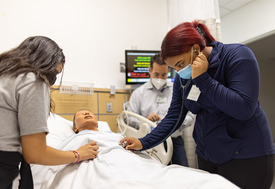 High school students use stethoscopes to hear body sounds from manikin in hospital simulation room