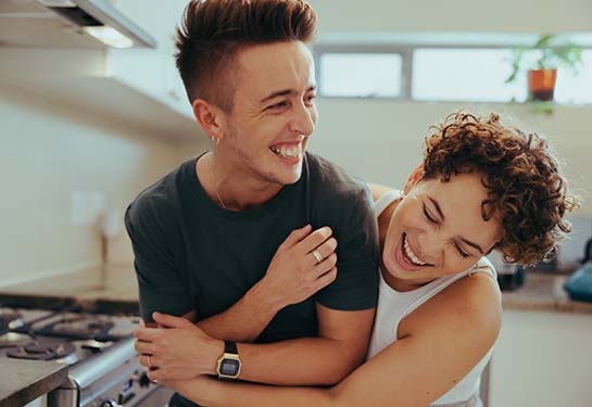 A young LGBTQIA+ couple laugh together in the kitchen.