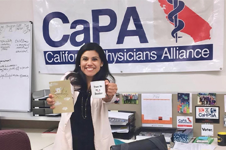 Angelica Martin wearing a white lab coat stands in her former office in front of a banner reading “California Physicians Alliance”
