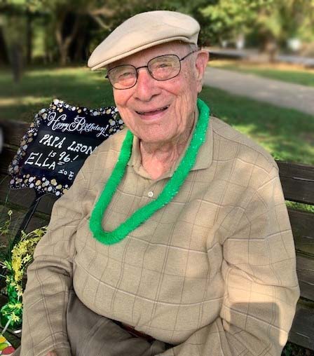 96-year-old man sits on a park bench, wearing a tan hat, glasses and a green lei around his neck. 