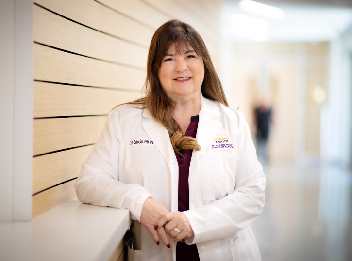 Deb Bakerjian stands in front of wooden wall wearing white lab coat and smiles