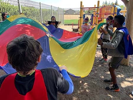 Children on a playground laugh while tossing a brightly colored parachute in the air. 