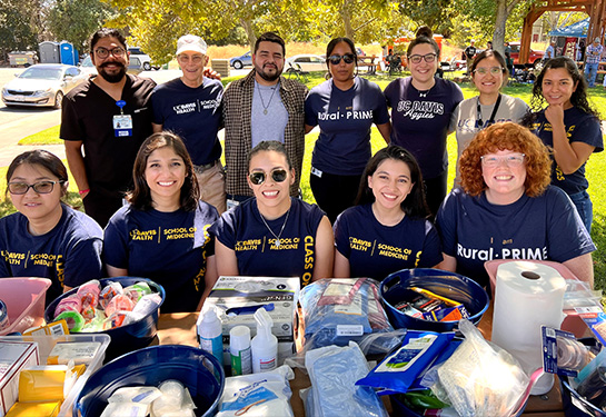 A group of 12 students wearing School of Medicine blue shirts pose before a picnic table with medical supplies such as bandages