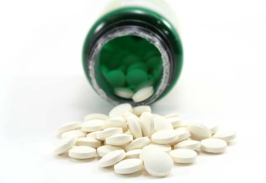 Round, off-white folic acid pills spill out from the mouth of a green vitamin bottle against a white 