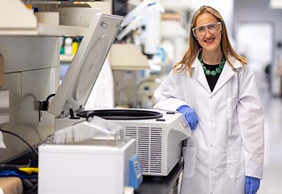 Associate Professor Bethany Cummings stands in her lab, wearing a white coat, next to equipment she uses to conduct metabolic research. 
