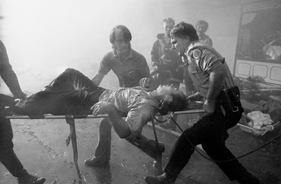 Two firefighters carry a stretcher with an injured man lying on it while bystanders look on