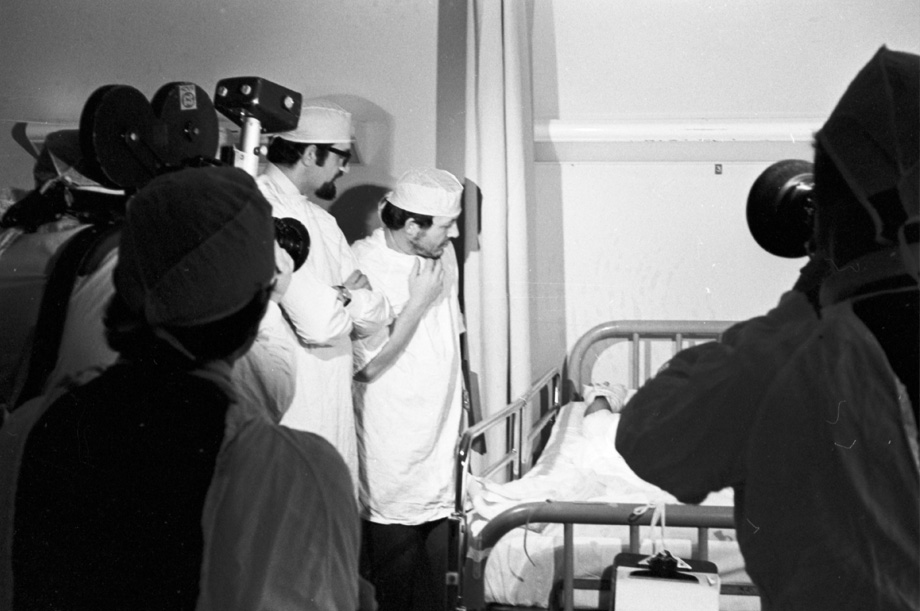 Doctors in background wearing white gowns standing over burn patient with TV video cameras in foreground