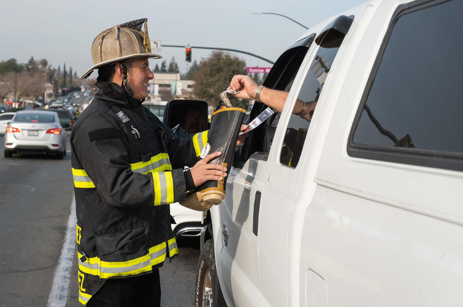 Firefighter in full gear stands outside a white truck holding fire boot while driver places dollar bill into boot