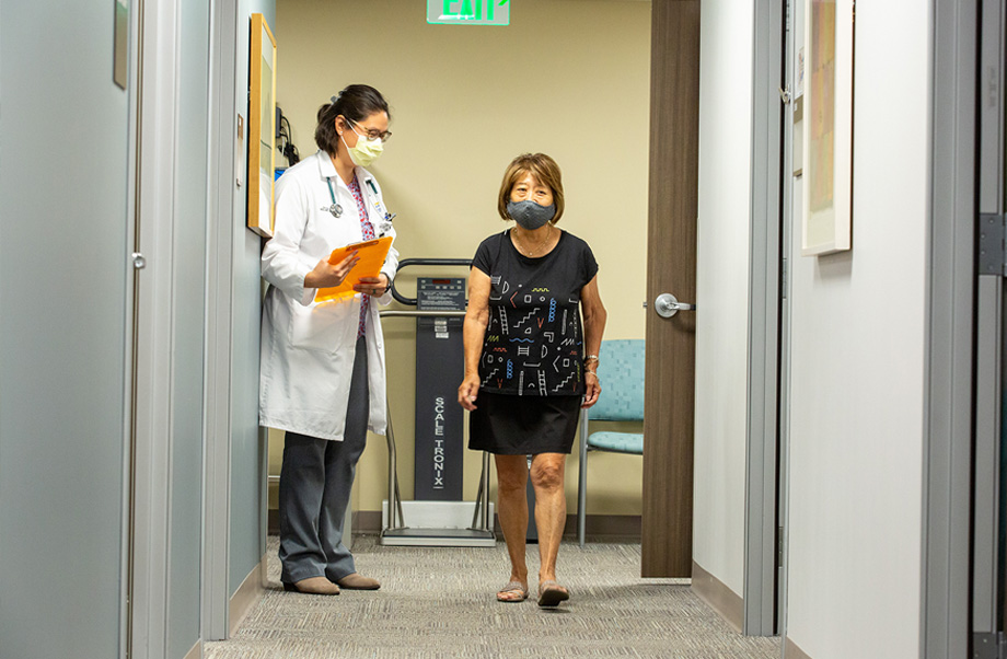 Nurse practitioner wearing white coat and holding clipboard watching female patient walk down hallway