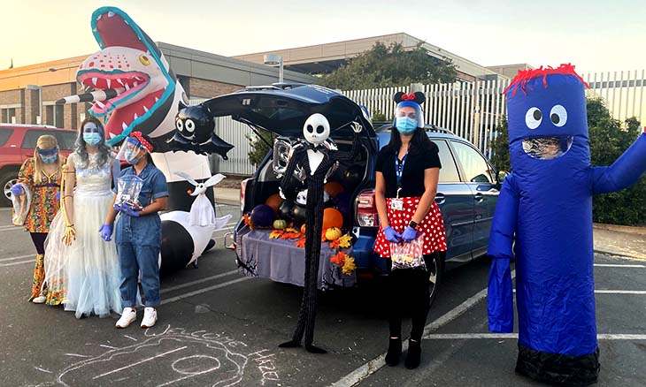 Five costumed people in front of an SUV with an open trunk decorated with pumpkins and leaves. Costumes include 70’s garb, a wedding dress, a nurse, Minnie Mouse and a blue inflatable monster.