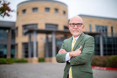 Portrait of a middle-aged bald man wearing an olive green suit, standing in front of a brown and glass building.