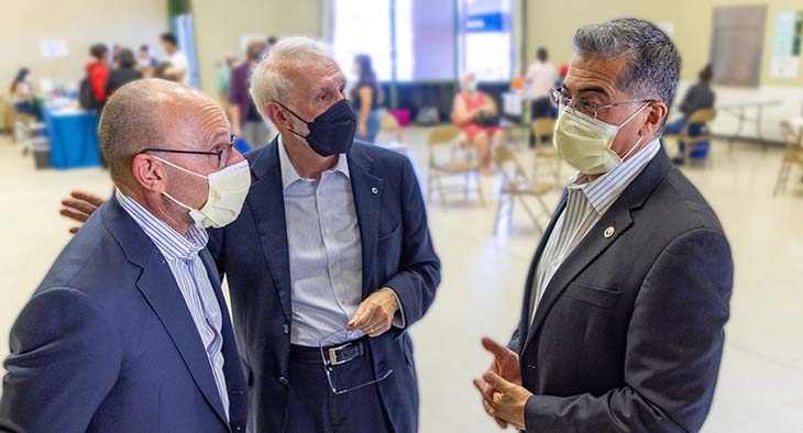 Three adult men wearing face masks gather in conversation while patients get vaccinations in the background.