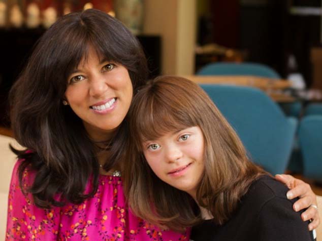 A woman with short black hair sits on a couch next to a young woman with Down syndrome. 