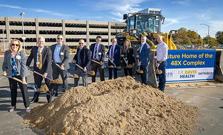 Pile of dirt with 9 individuals holding up shovels loaded with dirt, yellow front loader parked to the left