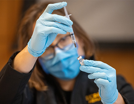 A UC Davis Health employee wearing blue gloves and blue face mask uses a syringe to extract vaccine from a small bottle 
