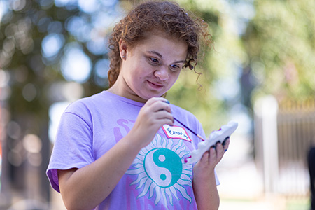 A teenage girl with brown curly hair wearing a light purple shirt with a yin and yang design stands painting an item she holds in her hand.