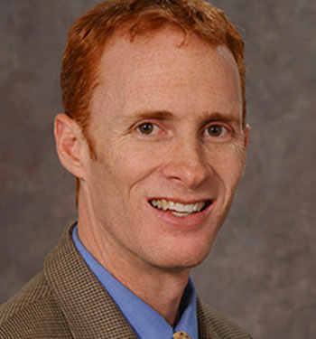 Man with red hair cut short, wearing blue shirt, yellow tie and plaid brown blazer on gray backdrop.