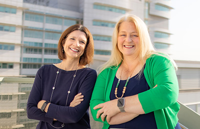 Two women stand next to a concrete structure, crossing their arms One wears a navy sweater, the other a navy dress and Kelly-green sweater. Both are smiling. UC Davis Medical Center is in the background.