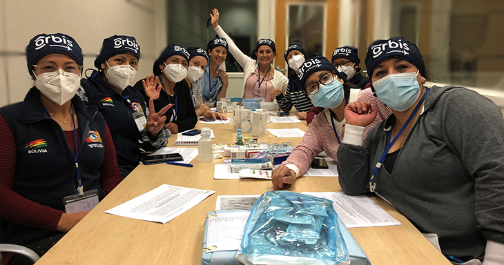 A group of women wearing surgical masks and blue hats with the word “Orbis” printed on them in white sit around a conference table.
