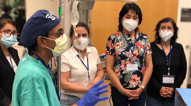 A man in green hospital scrubs and purple protective gloves, talks to a group of women in a hospital setting.  